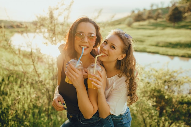 Las niñas felices se divierten, beben cócteles de vasos de plástico con paja en gafas de sol, vestidos con camisa blanca y negra, al atardecer, expresión facial positiva, al aire libre