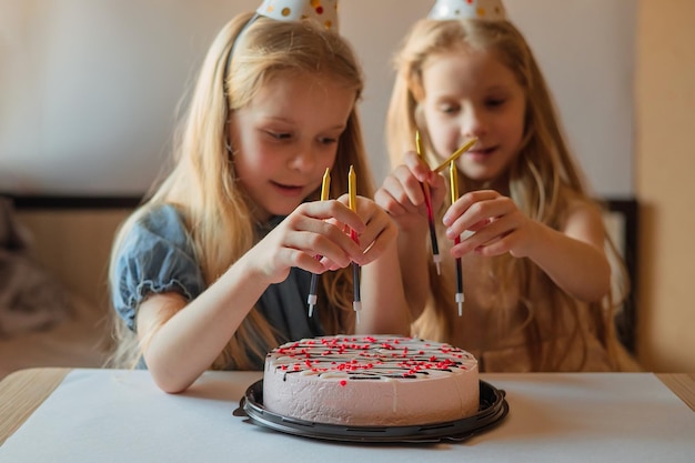 Niñas hermanas gemelas están felices de decorar pastel de cumpleaños con velas sombrero de vacaciones hecho en casa celebración de cumpleaños de niños fiesta interior durante el aislamiento de cuarentena por enfermedad