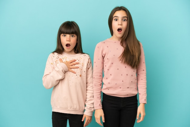 Niñas hermanas pequeñas aisladas sobre fondo azul con sorpresa y expresión facial conmocionada