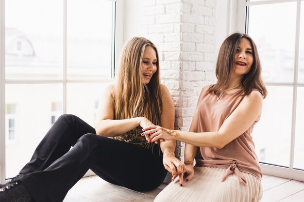 Niñas, hermanas sentadas en casa en la ventana, comunicación con un amigo, risas y diversión, patrón de henna en sus manos, tradiciones, personas, cuidado de la piel, familia, mujeres.