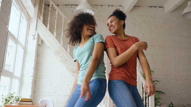 Niñas hermosas jóvenes de raza mixta bailando en una cama juntas divirtiéndose en el dormitorio en casa