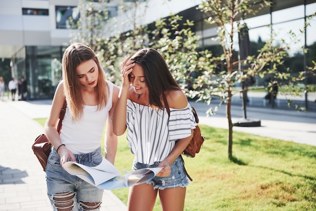 Niñas leyendo el mapa de la ciudad y buscando hotel. Los turistas encantadores con mochilas determinan el concepto de conocimiento del mundo.