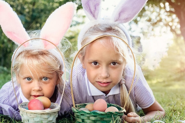 Foto niñas lindas con divertidas orejas de conejo recogen huevos de pascua en una canasta de mimbre sobre un césped contra un fondo verde borroso concepto de vacaciones de primavera