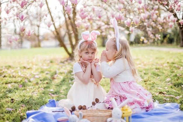 Las niñas lindas están sentadas en la hierba cerca de la mogolia. Chicas en disfraces de conejitos de Pascua. Primavera.