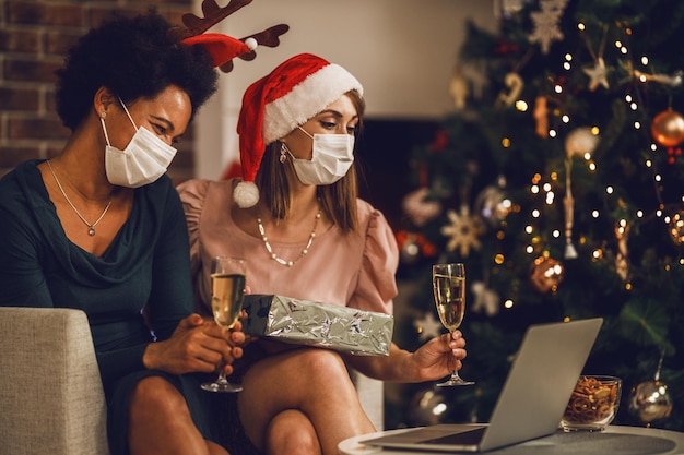 Niñas multiétnicas sonrientes con mascarilla sosteniendo una copa de champán y presentando una fiesta de Navidad o Año Nuevo en línea con amigos durante la pandemia.