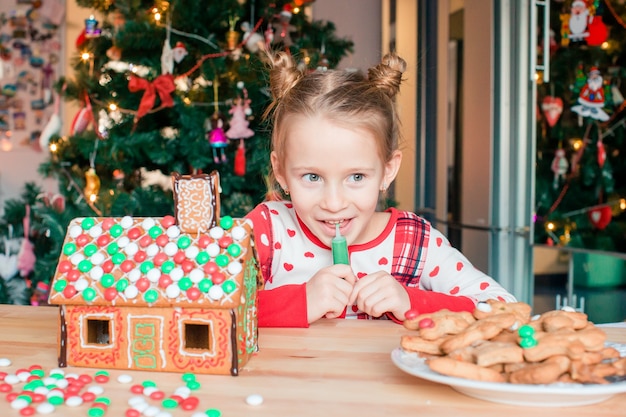 Niñas que hacen la casa de pan de jengibre de la Navidad en la chimenea en sala de estar adornada.