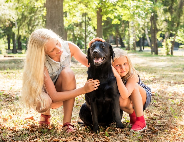 Niñas rubias se sientan abrazando a un perro negro en un parque