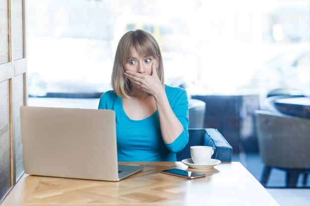 ¡De ninguna manera! Retrato de una joven increíble con el pelo rubio y una blusa azul sentada en un café, trabajando y haciendo una videollamada en una laptop, señalando con el dedo al compañero con la cara conmocionada, en el interior