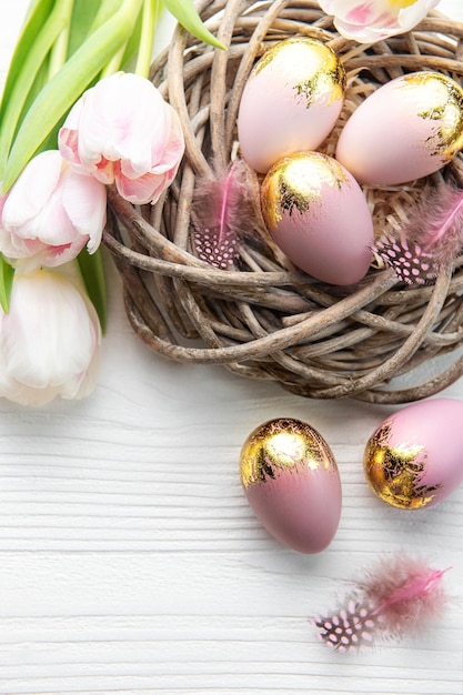 Ninho com ovos de Páscoa pintados de rosa e cores douradas sobre um fundo branco de madeira