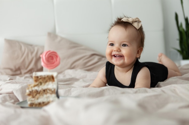 Una niñita divertida se arrastra en la cama hacia un delicioso pastel Cumpleaños