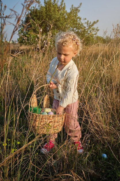 Una niñita divertida con suéter blanco recogiendo huevos de Pascua teñidos en una cesta de mimbre en una pradera verde cubierta de hierba en la naturaleza soleada de primavera