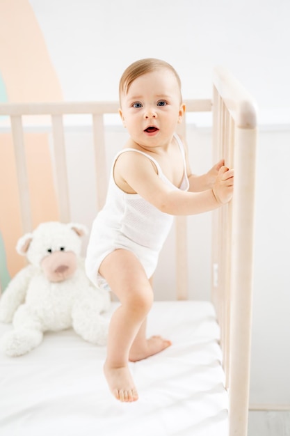 Una niñita está de pie en la cuna y sonriendo o riéndose en casa en la guardería con un estilo de vida de traje blanco