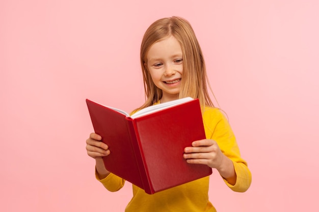 Una niñita inteligente y positiva que lee un gran libro rojo y sonríe felizmente aprendiendo la tarea siendo curiosa sobre el concepto de educación del conocimiento escolar tiro de estudio interior aislado en un fondo rosa
