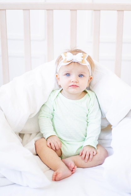 Una niñita linda y graciosa sentada en una cuna con una manta en una cama de algodón blanco con un traje verde en una habitación infantil luminosa con un lazo en la cabeza