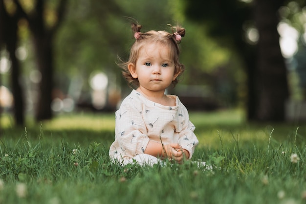 una niñita linda con un peinado divertido está sentada en un césped verde floreciente en el estilo de vida del parque