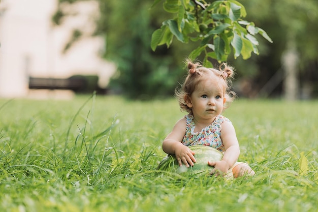 una niñita linda con un peinado divertido está sentada en un césped verde floreciente en el estilo de vida del parque