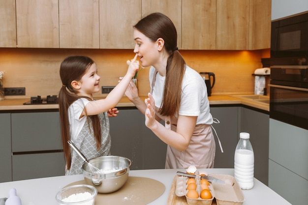 Una niñita linda y su joven madre con delantales juegan con harina y se ríen mientras amasan la masa en la cocina