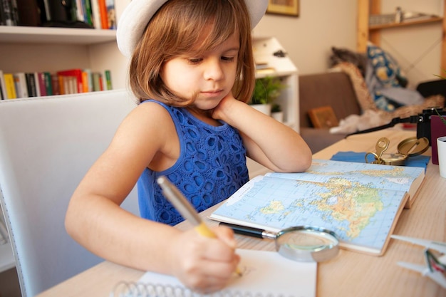 Una niñita de moda con ropa informal de verano y sombrero blanco está haciendo la tarea o escribiendo notas consultando un mapa geográfico pequeña viajera planeando un viaje alrededor del mundo con un explorador de la imaginación