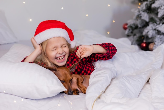 niñita rubia con sombrero de Papá Noel y pijamas está durmiendo en la cama con su amado perro salchicha