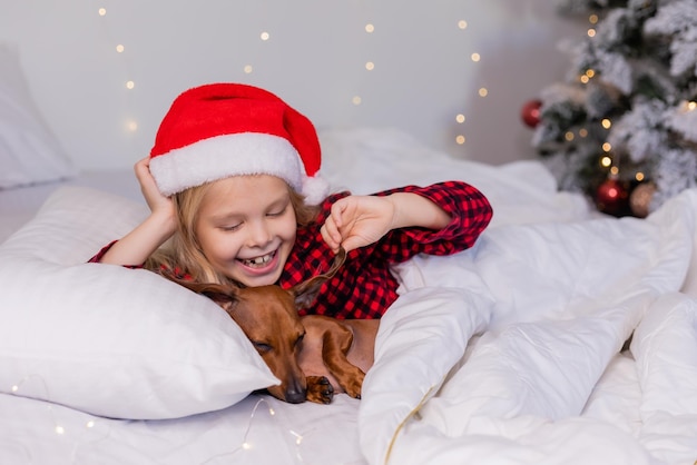 niñita rubia con sombrero de Papá Noel y pijamas está durmiendo en la cama con su amado perro salchicha