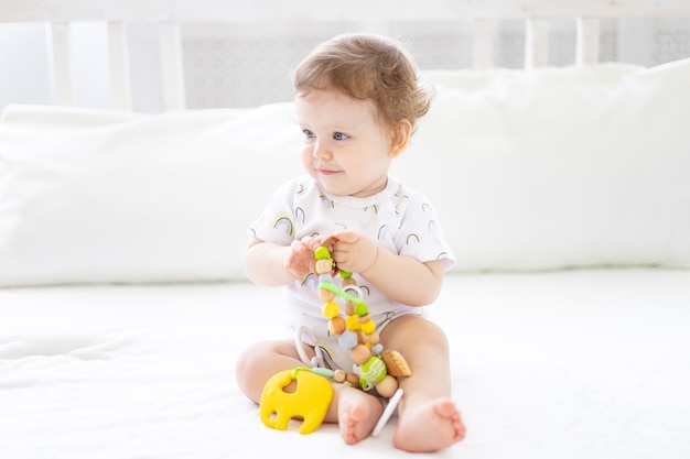 Una niñita sana y linda con un traje blanco está sentada en la cama con sábanas blancas sonriendo jugando con juguetes para el desarrollo temprano de los niños