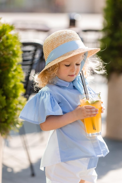 niñita con sombrero de paja y vestido azul bebe jugo de un vaso al aire libre