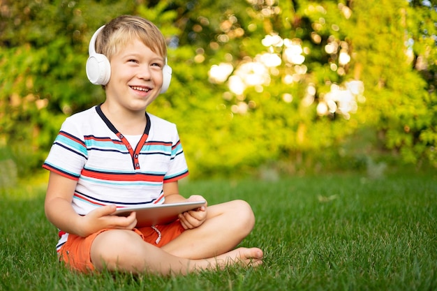 Un niñito feliz con auriculares inalámbricos sosteniendo una tableta al aire libre en un parque de verano sonriendo