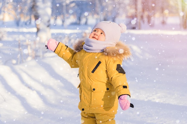 Niño de 12 a 17 meses en ropa de invierno amarilla mira los copos de nieve que caen mientras camina en un parque de invierno