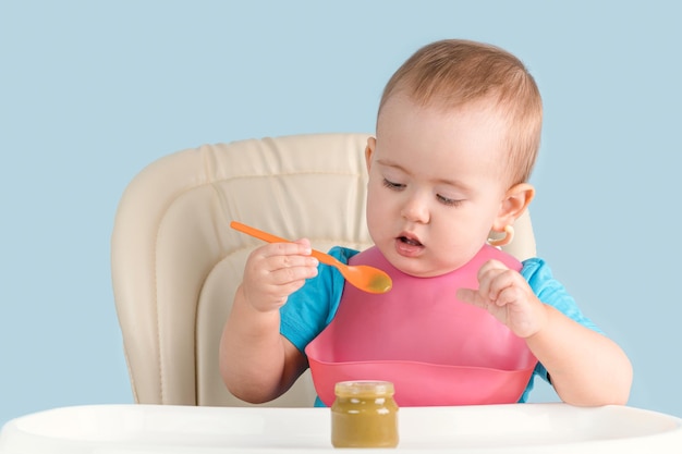 un niño de entre 12 y 23 meses se sienta en una silla de alimentación y aprende a comer puré de brócoli de un frasco de vidrio de comida para bebés con una cuchara por su cuenta