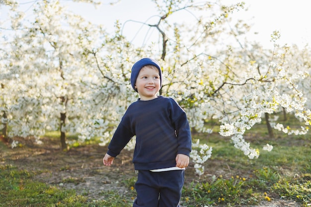 Un niño de 3 años con un suéter y un sombrero corre por un jardín floreciente Ropa para niños de 3 años Un niño eotiano feliz es blanco entre manzanos en flor