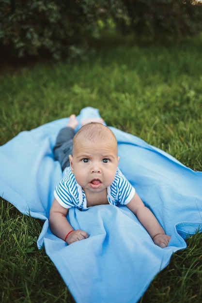 Niño de 3 meses yace boca abajo sobre una colcha azul en el parque alrededor de la hierba verde y los árboles. Las emociones de alegría de los niños