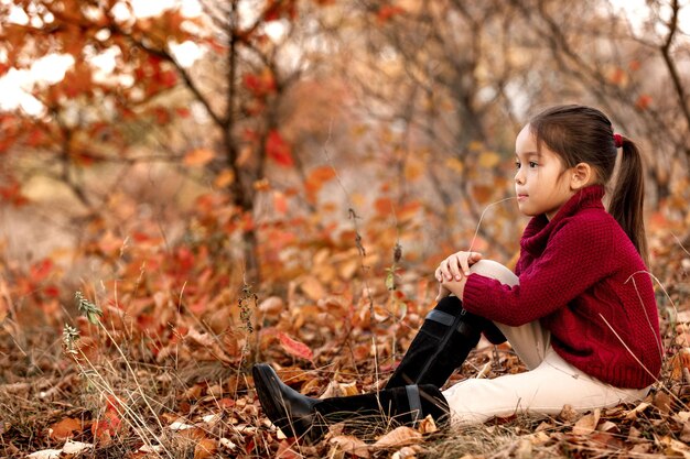 Niño de 5 años de moda posando en el parque de otoño. niña sentada en hojas caídas