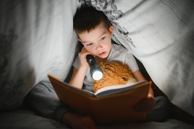Un niño de 56 años está leyendo un libro por la noche en la oscuridad debajo de una manta con un oso de juguete.