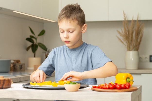 Niño 7-10 en camiseta cocinando pizza en la cocina. Niño arregla los ingredientes en la base de la pizza