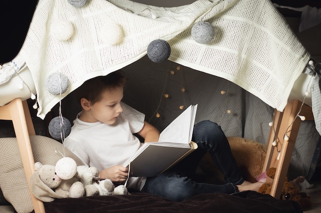 El niño de 7 a 11 años se sienta en una cabaña de sillas y mantas. Niño, libro de lectura en casa