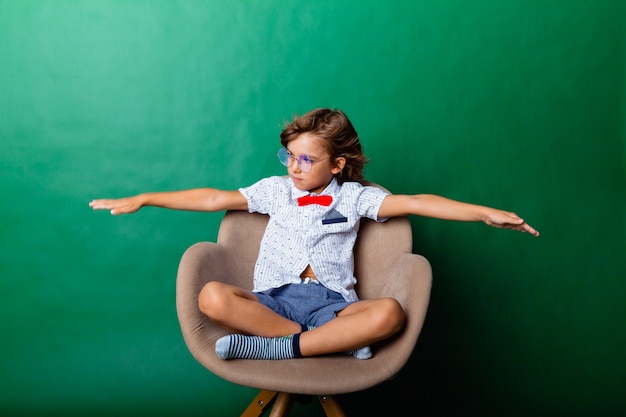 El niño de 7 años se sienta en una silla y levantó las manos por encima de la cabeza, aislado en un fondo verde. retrato de un chico guapo