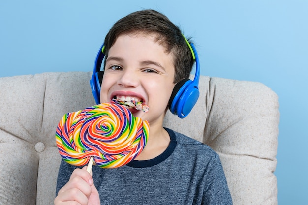 Foto niño de 8 años, con auriculares, mordiendo una paleta de colores y mirando a la cámara.