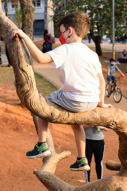 Niño de 8 años, con máscara, sentado en el tronco de un árbol en una plaza pública.