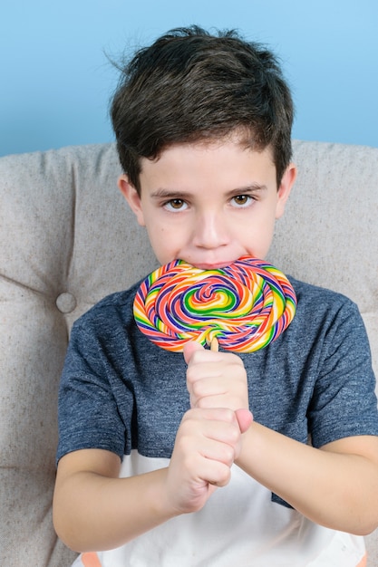 Niño de 8 años mordiendo una gran paleta de colores y mirando a la cámara (foto vertical).