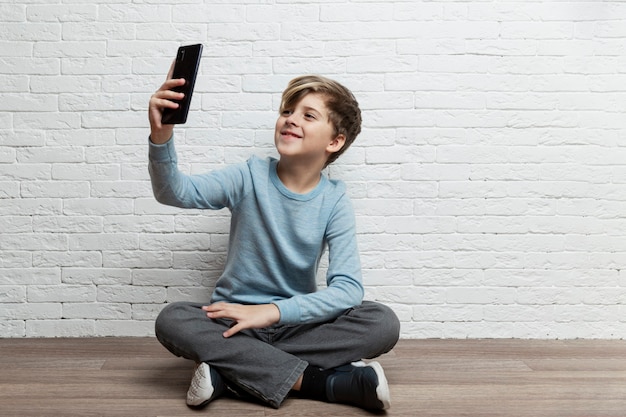 Un niño de 9 a 10 años se sienta con un teléfono en sus manos sobre un fondo de una pared de ladrillo blanco. Comunicación online y redes sociales. Espacio para texto.