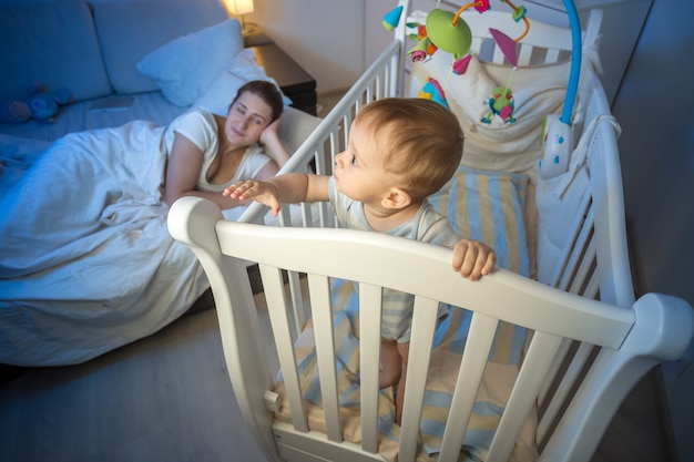 Niño de 9 meses de edad de pie en la cuna y despertando a su madre dormida