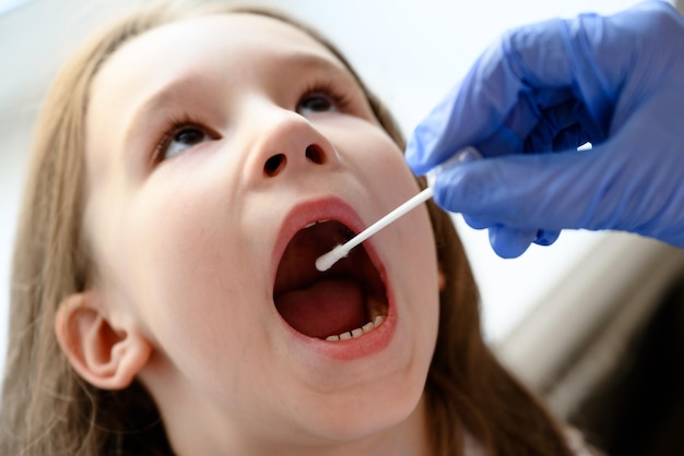 El niño abre la boca para la prueba de COVID19 El médico sostiene un hisopo para la muestra de saliva de un niño lindo