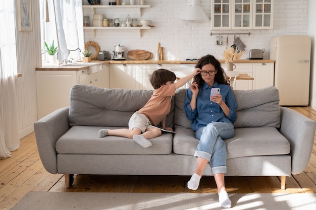 Niño aburrido molestar a la madre molesta empresaria leyendo el mensaje en el teléfono inteligente necesita la atención de mamá