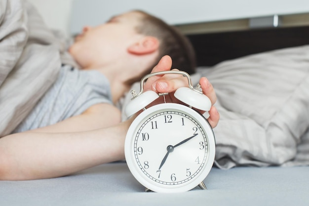 Niño acostado en la cama y deteniendo el despertador por la mañana