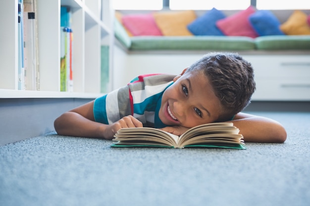 Niño acostado en el piso y leyendo un libro en la biblioteca