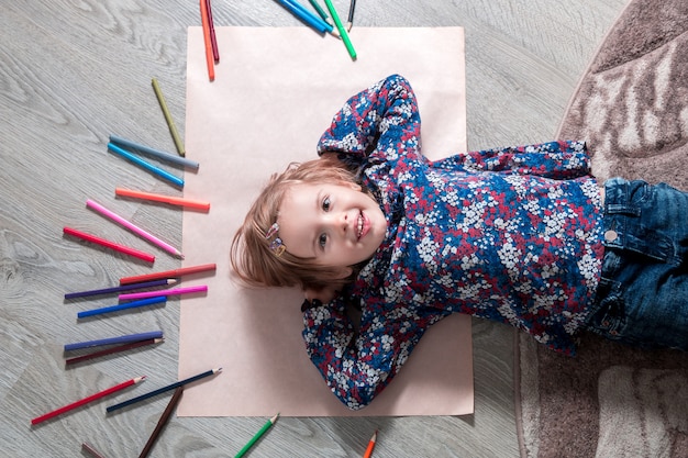 Niño acostado en el piso sobre papel mirando a la cámara cerca de lápices de colores. Pintando dibujando.