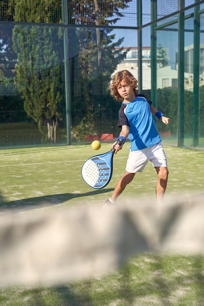 Niño activo en ropa deportiva golpeando la pelota con raqueta profesional mientras juega al pádel en el patio de recreo con red en el día de verano