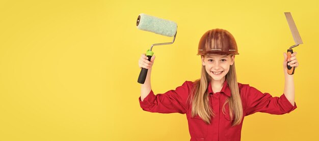 Niño adolescente alegre en casco de constructor con rodillo de pintura y espátula en pared amarilla Constructor de niños en diseño de cartel horizontal de casco Espacio de copia de encabezado de pancarta