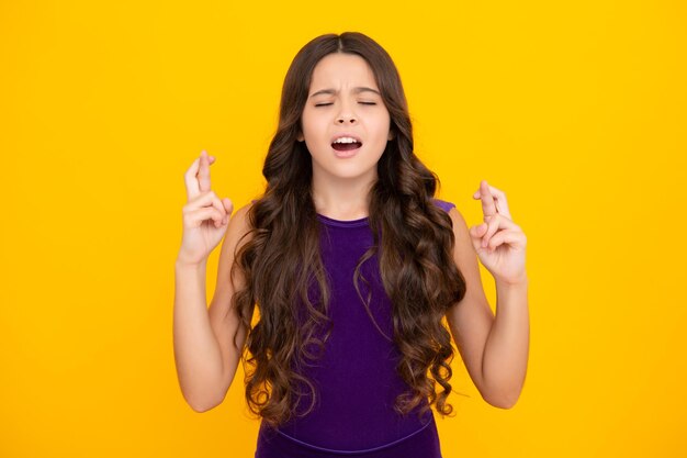 Foto niño adolescente con los dedos cruzados para la buena suerte jovencita reza y espera que los sueños se hagan realidad aislado sobre fondo amarillo