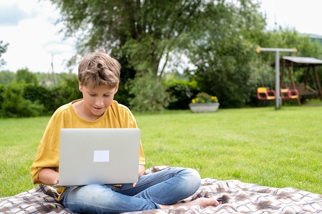 Niño adolescente escribiendo en su computadora portátil en el parque Niño estudiando o jugando a juegos de computadora al aire libre en el prado